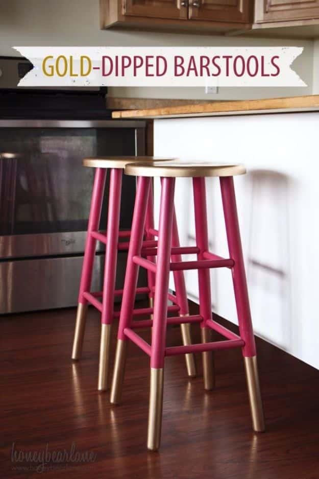 Gold dipped bar stools with raspberry pink legs and gold sprayed feet and seats, beside a stainless oven on a wood floor