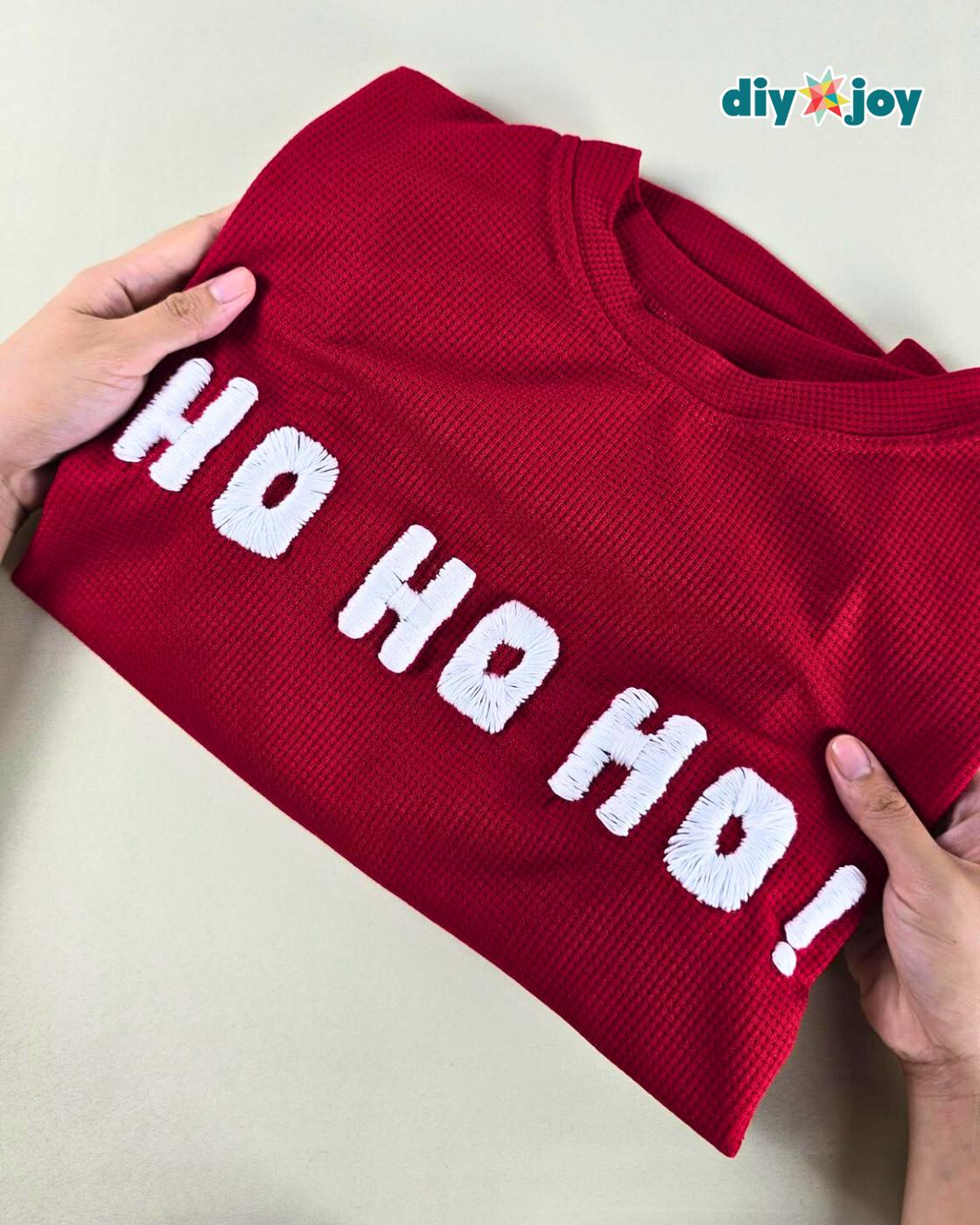 Christmas Sweater - two hands holding open a folded red waffle knit sweater with HO HO HO stitched across the front in raised white lettering