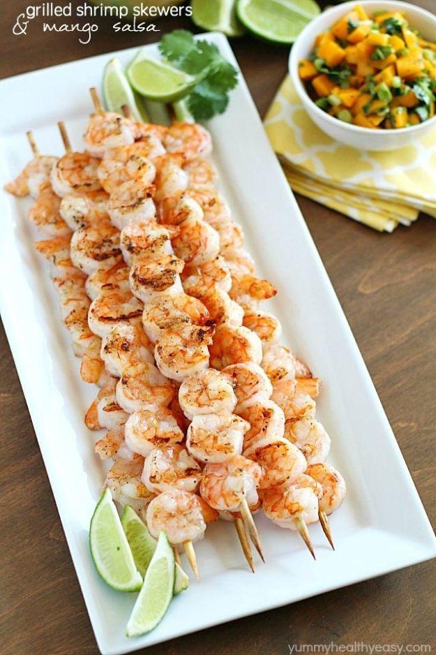 Grilled shrimp skewers lined up on a white platter with lime wedges and a bowl of chunky fresh mango salsa