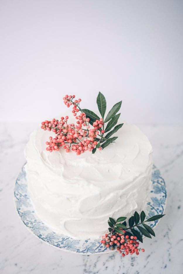 Small white frosted layer cake topped with tiny pink blossoms and green leaves on a pale blue cake stand