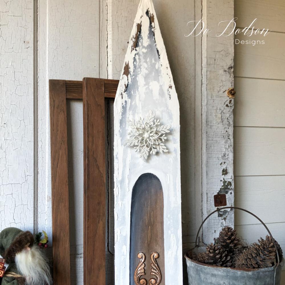Tall wooden church cut from a single board, painted white over brown stain with textured snow on the roof, beside pine cones