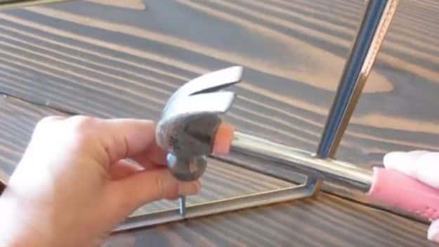 Close up of a hand holding a hammer above a slim black picture frame being assembled on a wooden work surface