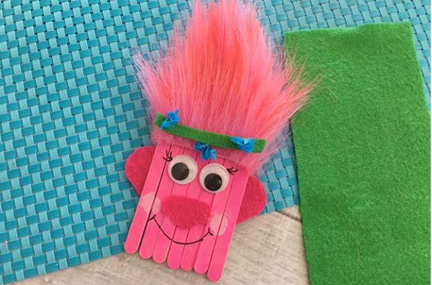 Popsicle stick Trolls Poppy with pink faux fur hair, felt ears and a green flower headband on a woven placemat