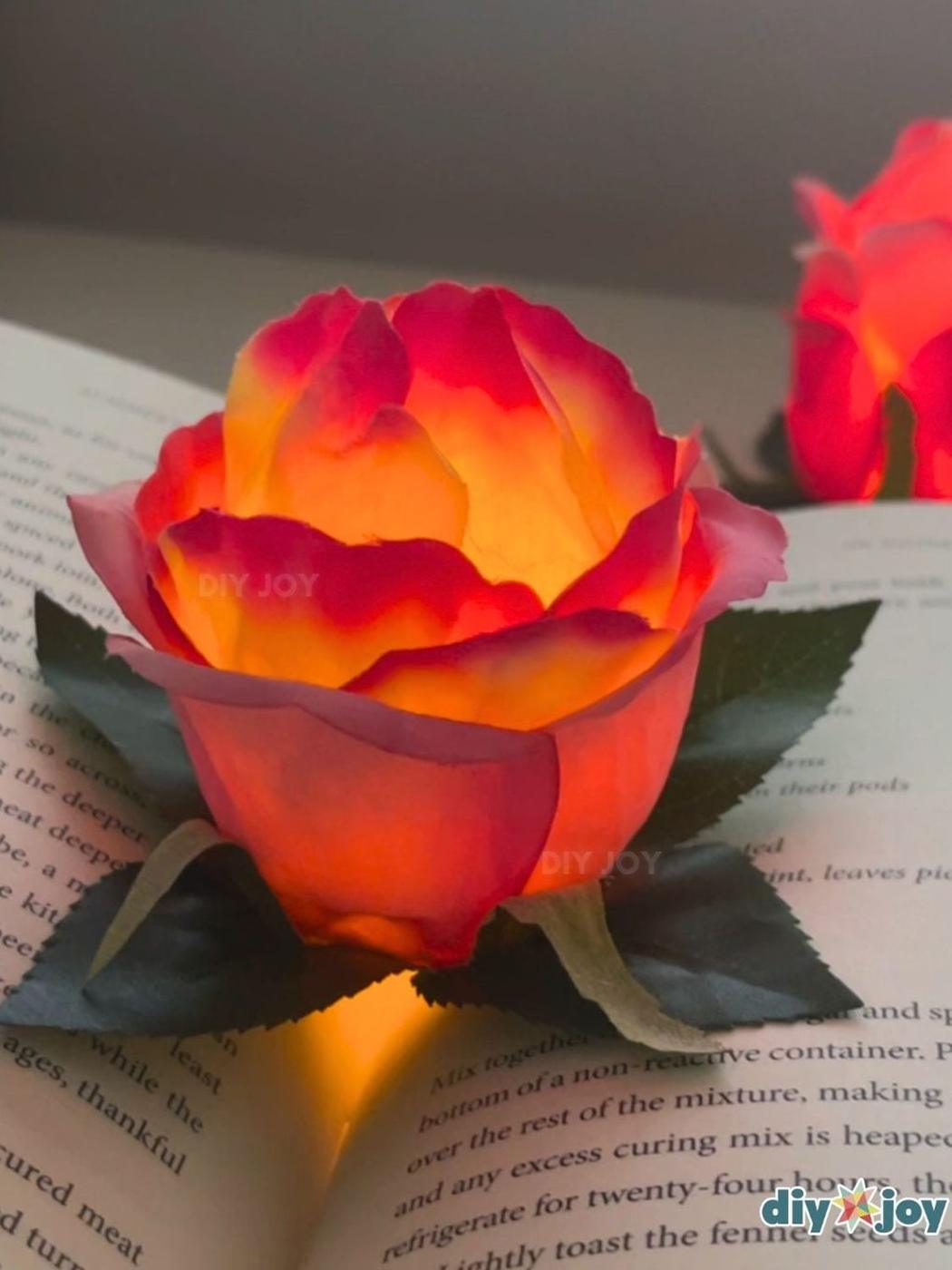 A red and gold faux rose glowing from a tea light inside its petals on an open book, with a second glowing rose behind