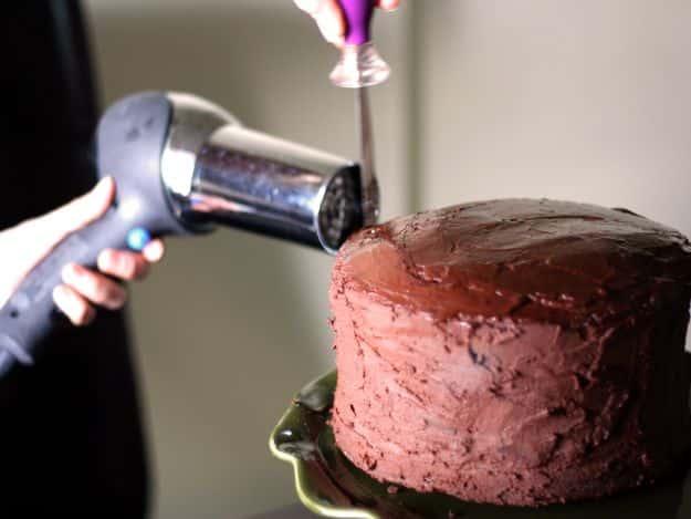 A hairdryer being held against the side of a chocolate ganache covered cake to smooth the frosting ridges