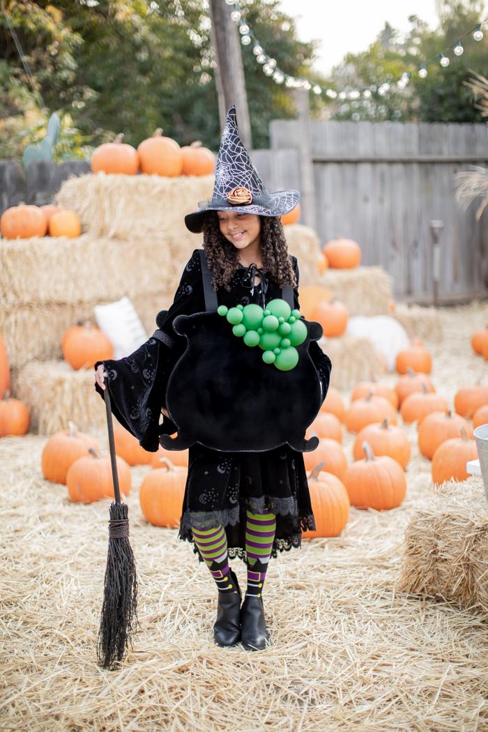 Child dressed as a witch wearing a black cauldron costume with green bubbles and holding a broom in a pumpkin patch