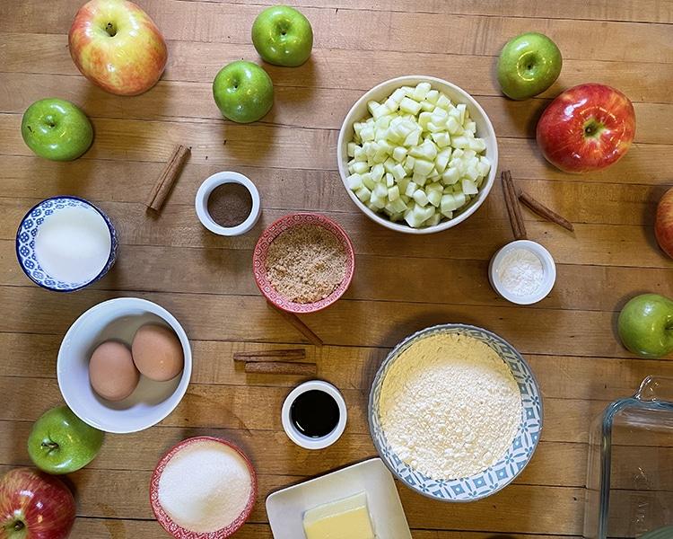 Easy Apple Recipes - ingredients arranged in a circle on wood, including whole apples, diced apple, eggs, brown sugar, flour, butter, cinnamon sticks, vanilla, milk and a glass bak