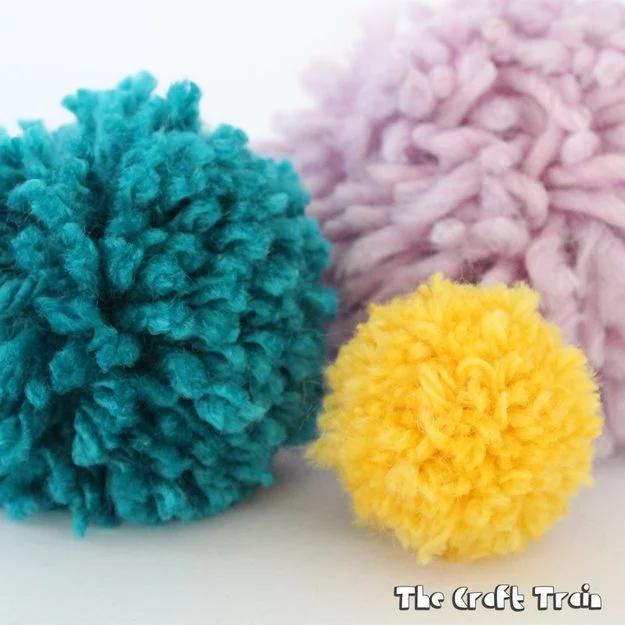 DIY cardboard pom pom maker, three yarn pom poms in teal, pale pink and yellow sitting on a white surface
