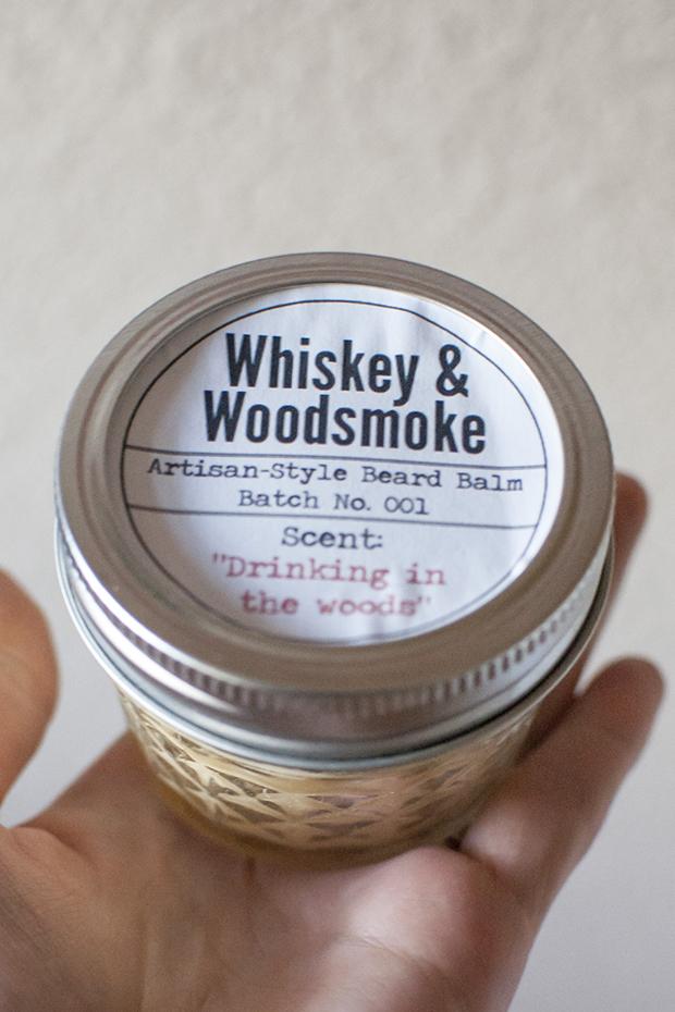 A hand holding a small mason jar of homemade beard balm with a printed Whiskey and Woodsmoke label on the lid