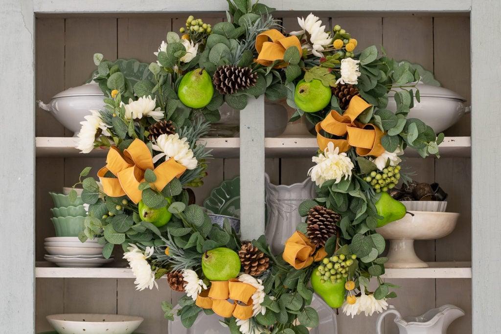 Wreath of green pears, pinecones, white daisies and mustard ribbon displayed on a hutch for the autumn season