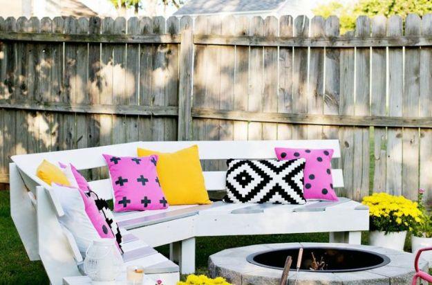 Curved fire pit bench painted white with bright pink and yellow cushions around a stone fire pit by a fence