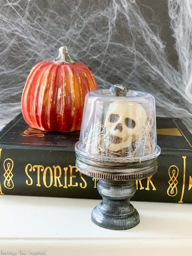 Miniature skull and Spanish moss sit under an inverted glass jar on a black and silver painted pedestal
