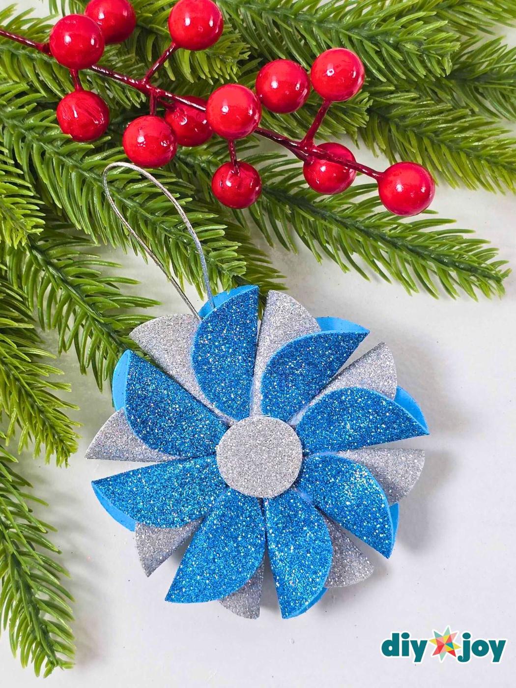 A pinwheel ornament of blue and silver glitter foam petals with a silver center hanging on a silver cord beside red berries