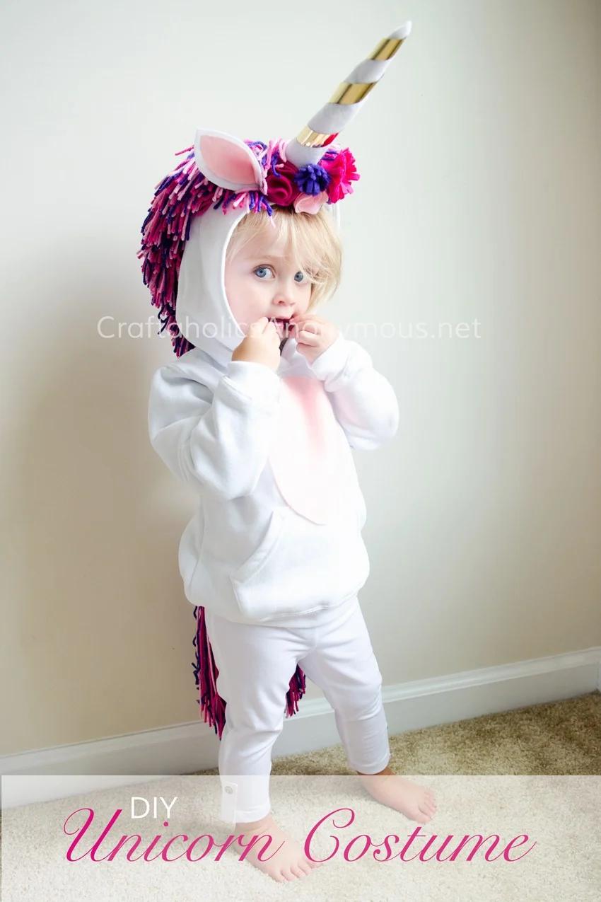 Child wearing a white unicorn hoodie costume with felt horn and ears, pink-purple yarn mane and source watermark