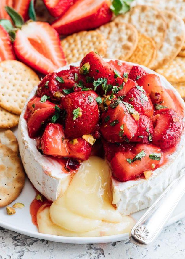 Roasted strawberry baked brie topped with honey roasted berries, chopped pistachios and mint beside crackers