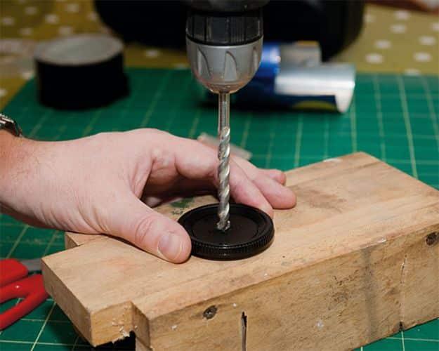 Pinhole camera conversion, a hand steadying a black camera body cap on wood while a drill bit cuts the center
