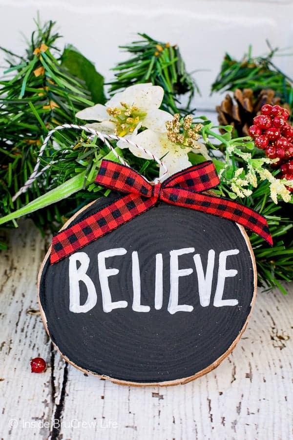 A black wood slice ornament lettered BELIEVE in white with a red buffalo check bow, propped against greenery and red berries