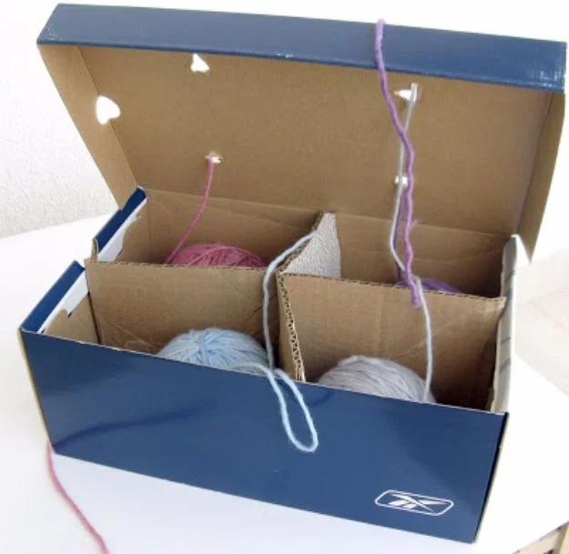 Open navy shoe box with cardboard dividers holding balls of yarn, strands threaded up through holes in the lid