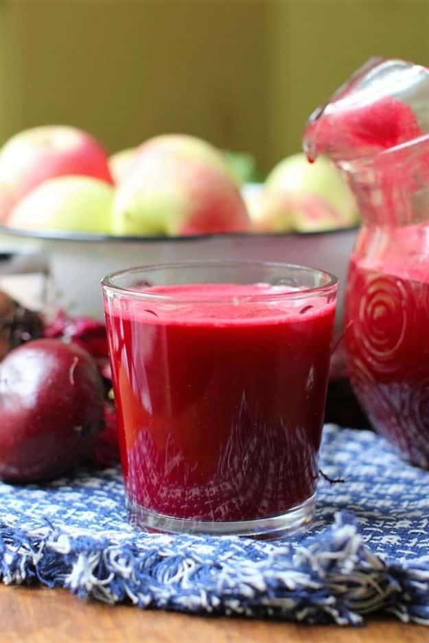 Apple beet juice in a glass on a blue cloth with a full pitcher, a bowl of apples and raw beets behind it