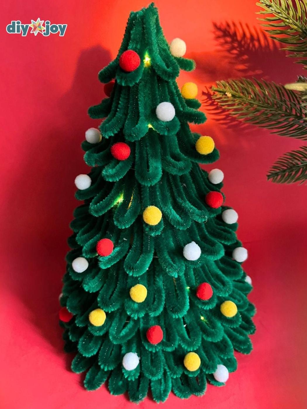A tabletop Christmas tree made of looped green pipe cleaners with red, yellow and white pom pom ornaments, lit from inside