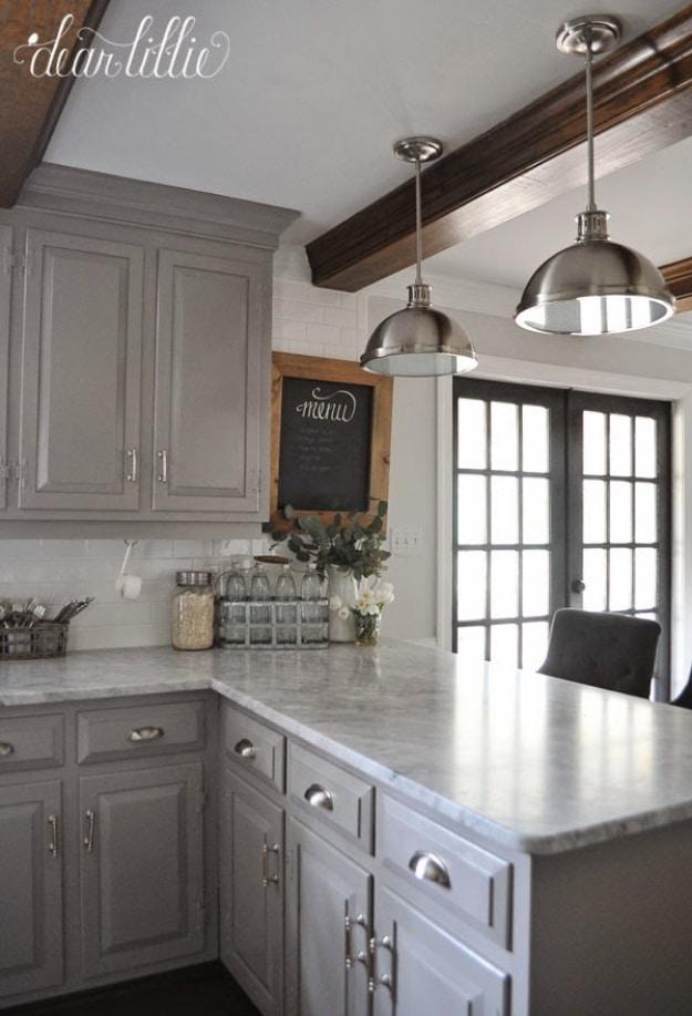 Grey painted kitchen cabinets with nickel cup pulls, marbled counters, two dome pendant lights and stained wood ceiling beams