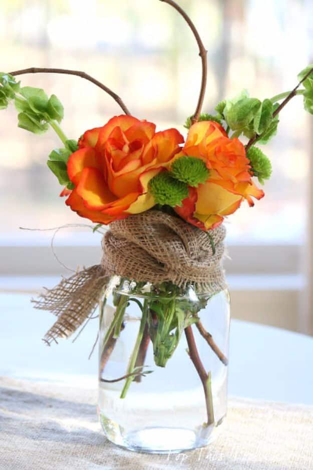 Mason jar centerpiece of orange roses and green blooms with a burlap tie and bare twigs arching above