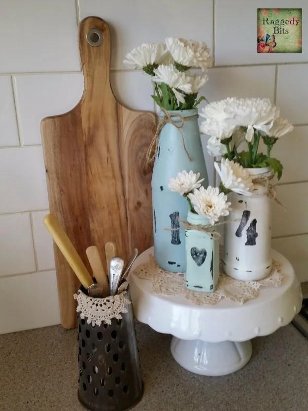 DIY homey vases, painted white and teal jars wrapped with twine and heart tags holding white daisies on a cake stand