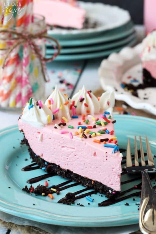 Slice of pink no bake strawberry cheesecake on a teal plate with a dark Oreo crust, sprinkles and whipped cream