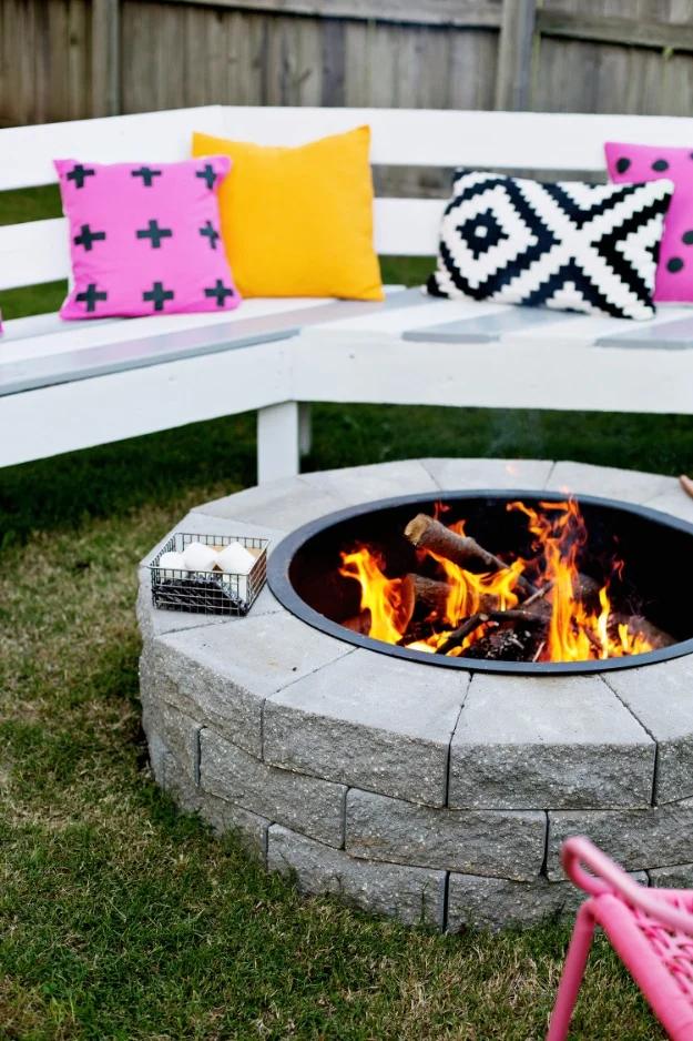 Stacked stone DIY firepit ring built in four steps, burning logs, with a white bench and bright pillows around it