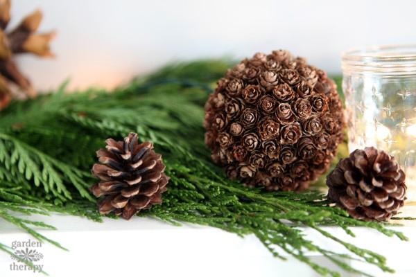 Pinecone sphere made of small cones glued over a ball, resting on cedar greenery beside loose pinecones