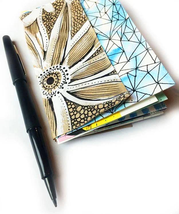 Two easy handmade art journals with painted covers, a gold and black drawn flower and a blue geometric web, and a brush pen