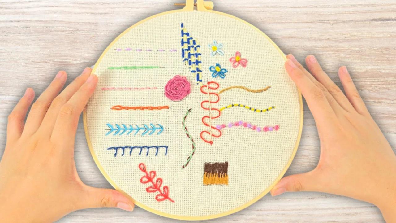 Two hands holding a yellow embroidery hoop of cream aida stitched with fifteen sample stitches in bright floss colors