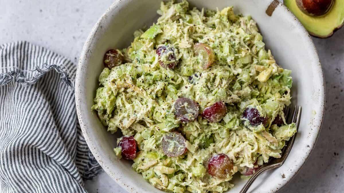 Avocado chicken salad with shredded chicken, red grapes and celery in a pale bowl with a striped cloth