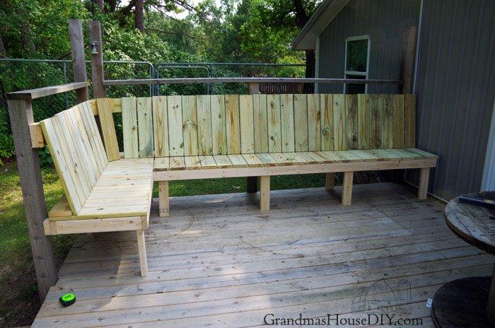 Wraparound corner deck bench in pale treated lumber running along two sides of a wooden deck by a gray house