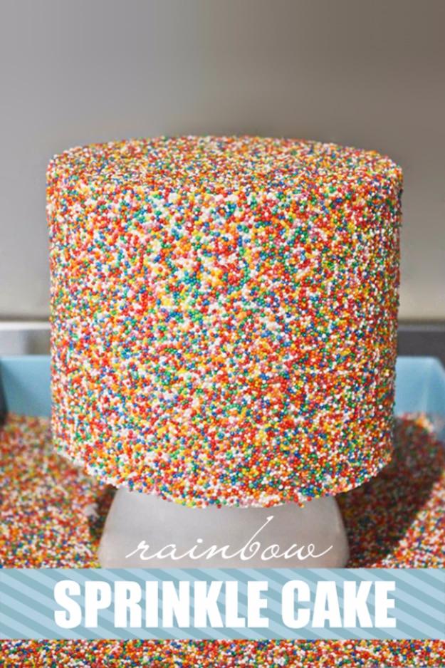 Tall birthday cake covered completely in rainbow sprinkles on a white cake stand against a blue wall