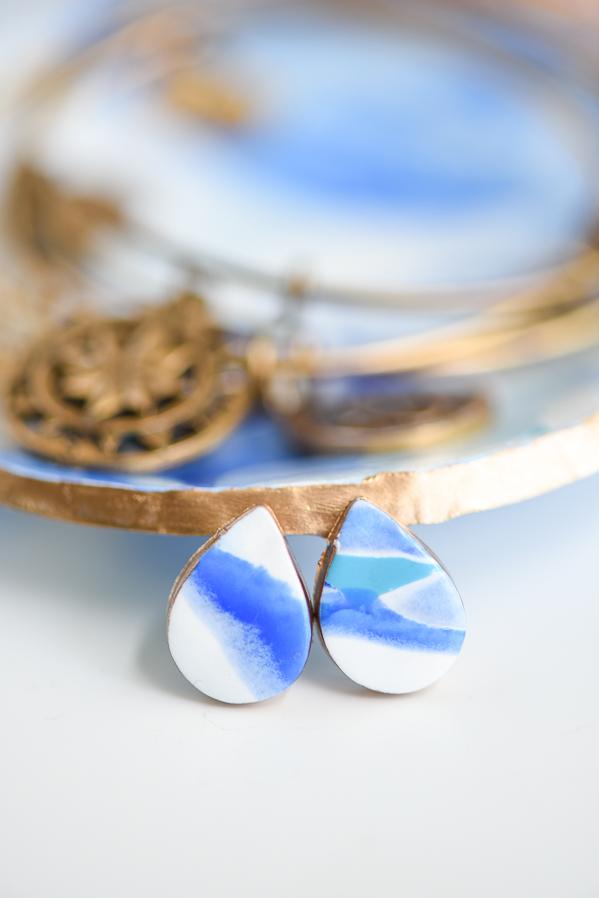 Marbled clay stud earrings, two blue and white teardrop studs with gold top edges beside a gold trinket dish
