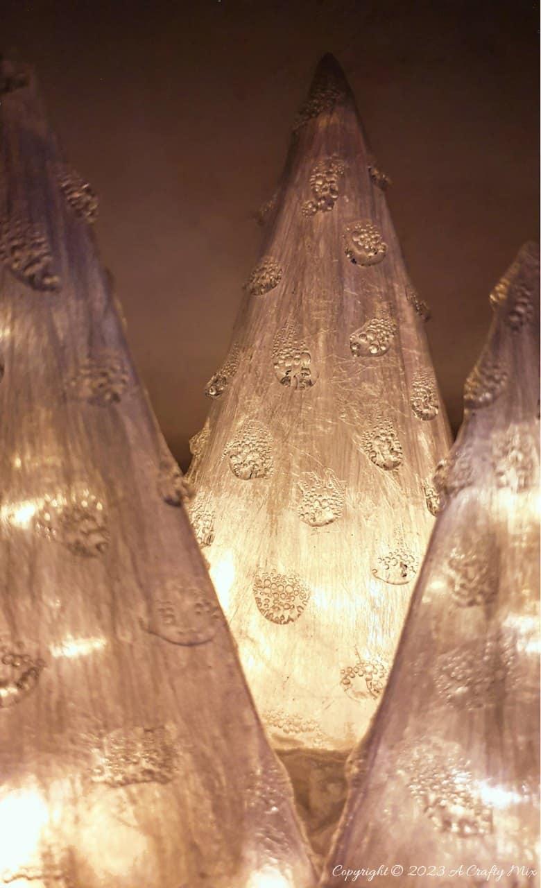 Three translucent cone Christmas trees made from layered cling wrap waste, glowing from fairy lights inside on a dark table