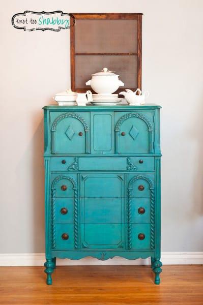 Ornately carved antique dresser painted peacock blue with a white tureen on top, standing on a wood floor