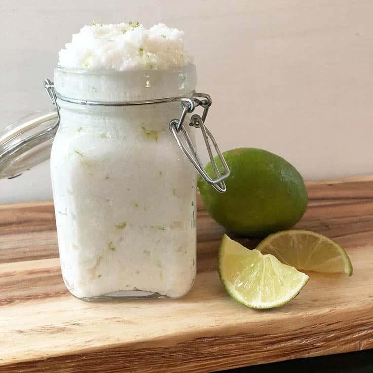 Square clip top glass jar filled with white coconut lime sugar scrub on a wood board beside fresh lime wedges