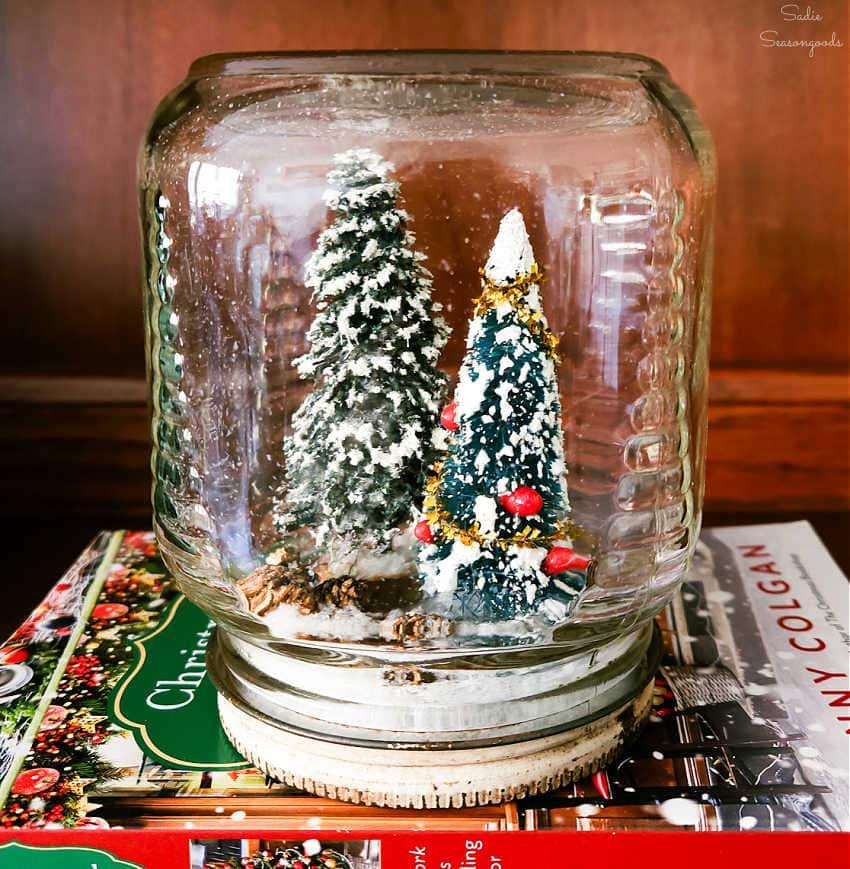 Vintage glass jar inverted over a snowy scene of two bottle brush Christmas trees, standing on stacked books