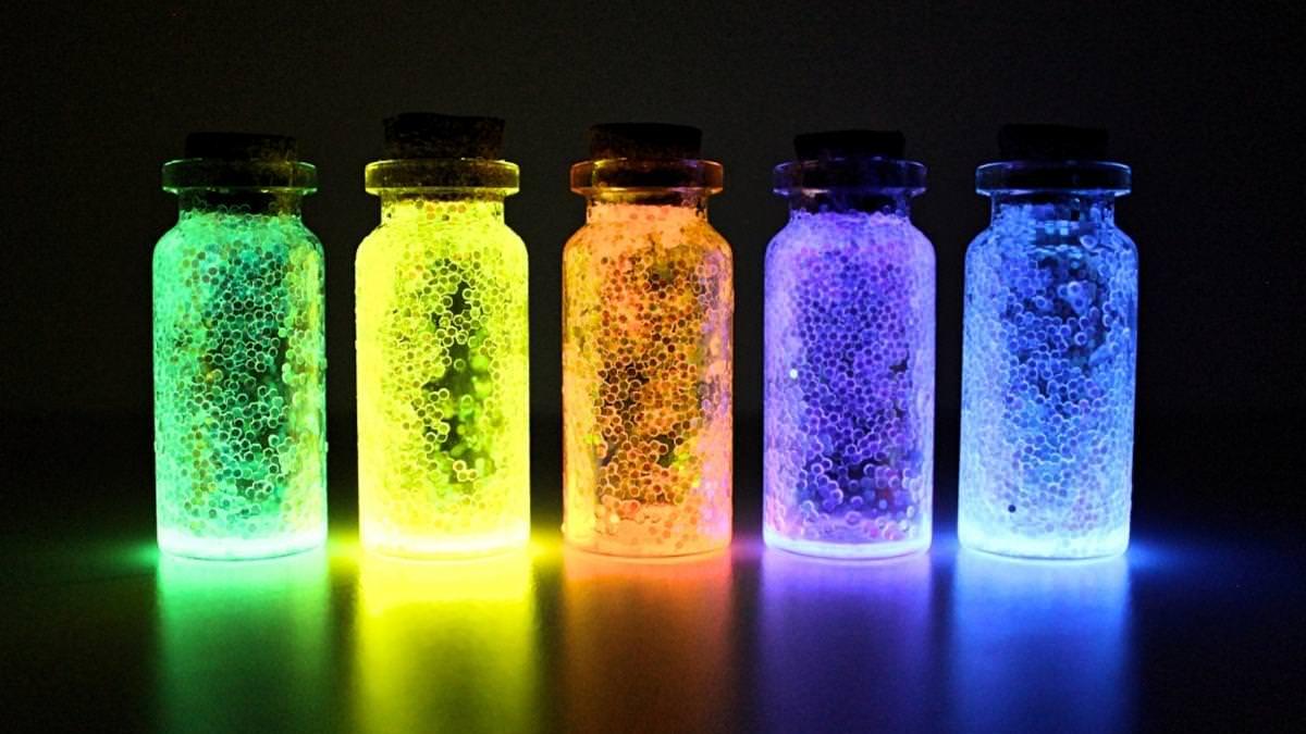 Five small corked bottles glowing green, yellow, orange, purple and blue with glitter inside in a dark room