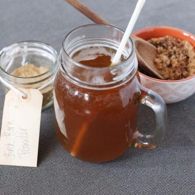 Homemade birch beer in a tall glass over ice with pieces of dried birch bark on the counter beside it