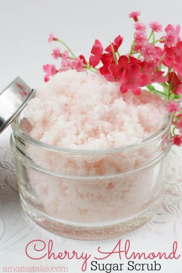 DIY cherry almond sugar scrub, a soft pink sugar scrub in a clear glass bowl with pink blossoms behind it