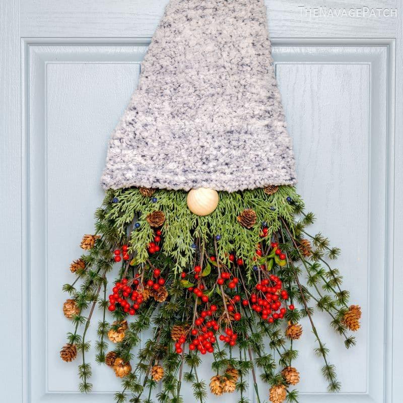 A gnome wreath on a blue door with a gray knit hat, a wooden bead nose and a beard of greenery, red berries and pinecones