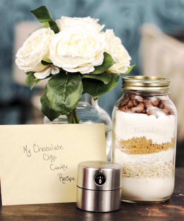 Mason jar of layered chocolate chip cookie mix beside a handwritten recipe card and a vase of white roses