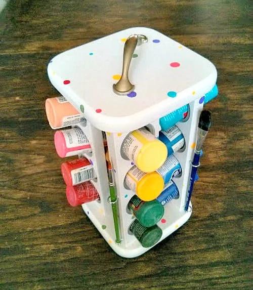 White spice rack spotted with colored dots holding craft paint bottles, with brushes in eye hooks and a handle