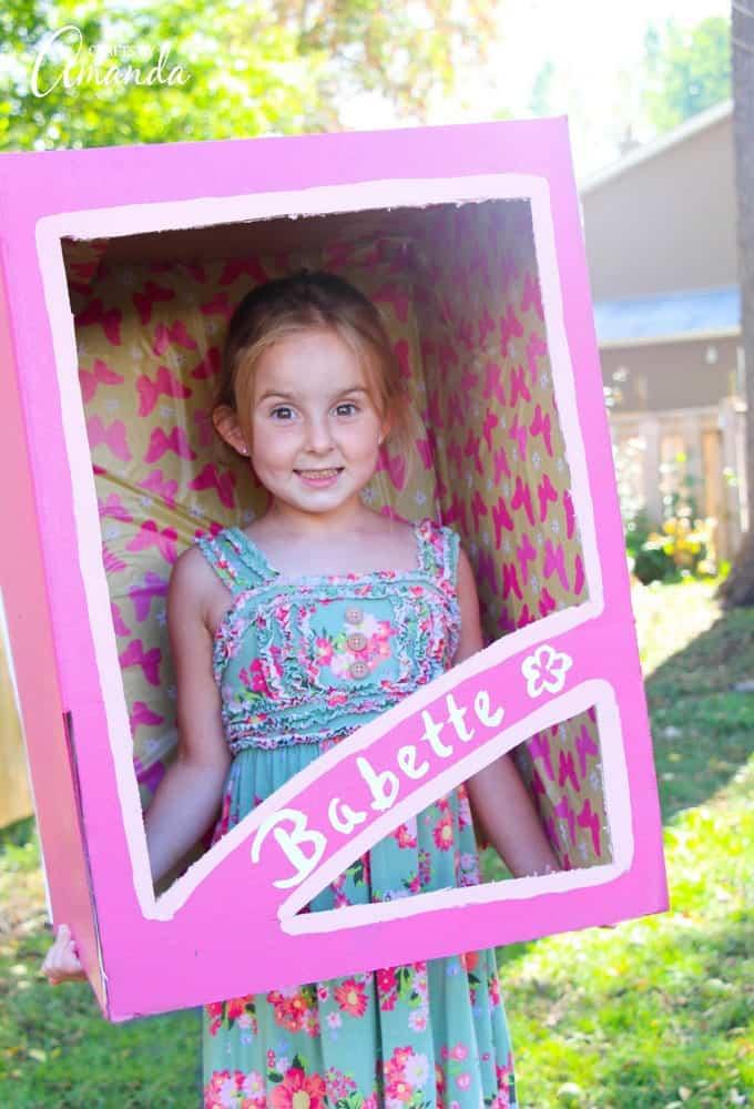 Child wearing a pink doll box costume made from cardboard with painted trim and patterned paper lining