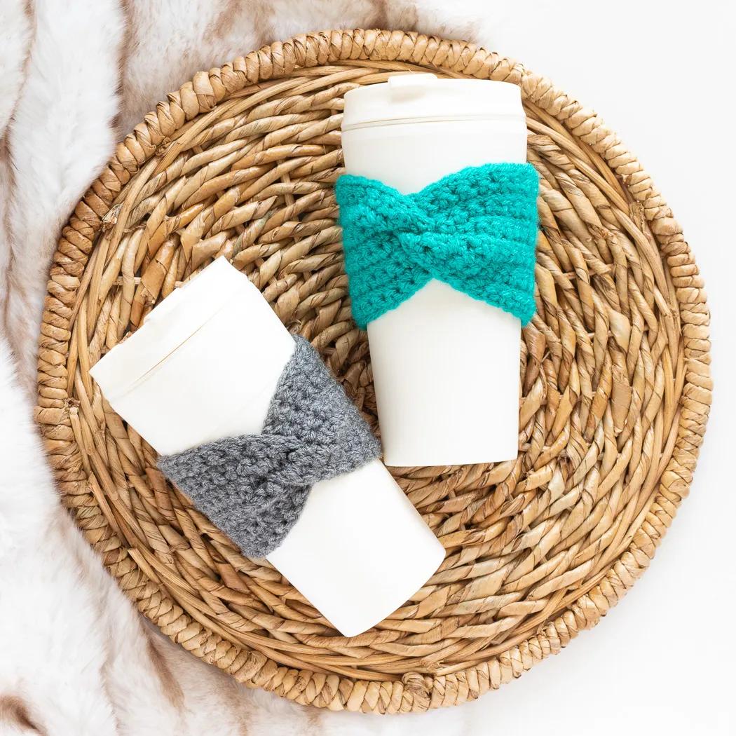 Two crochet cup cozies with a twisted bow front in teal and gray on white paper coffee cups on a woven placemat