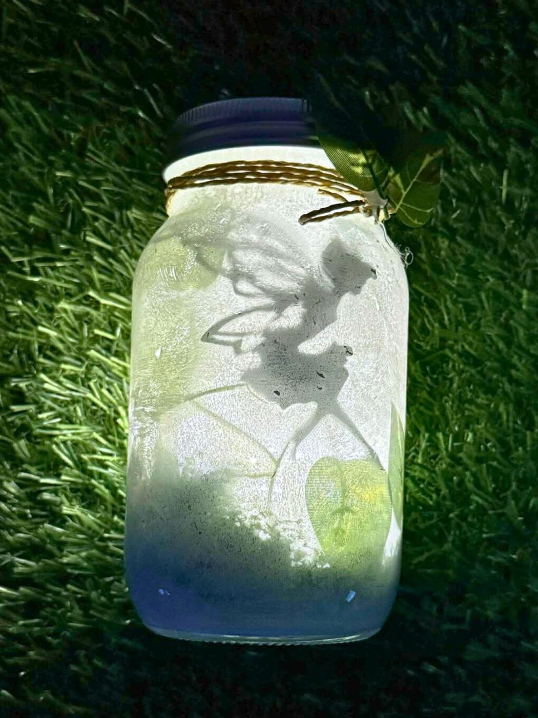 A frosted mason jar lantern glowing pale blue and white with a fairy silhouette inside, tied with twine and a leaf on grass