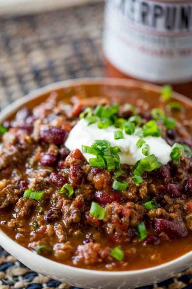 Slow cooker beef chili in a white bowl with sour cream and sliced green onions beside a beer growler